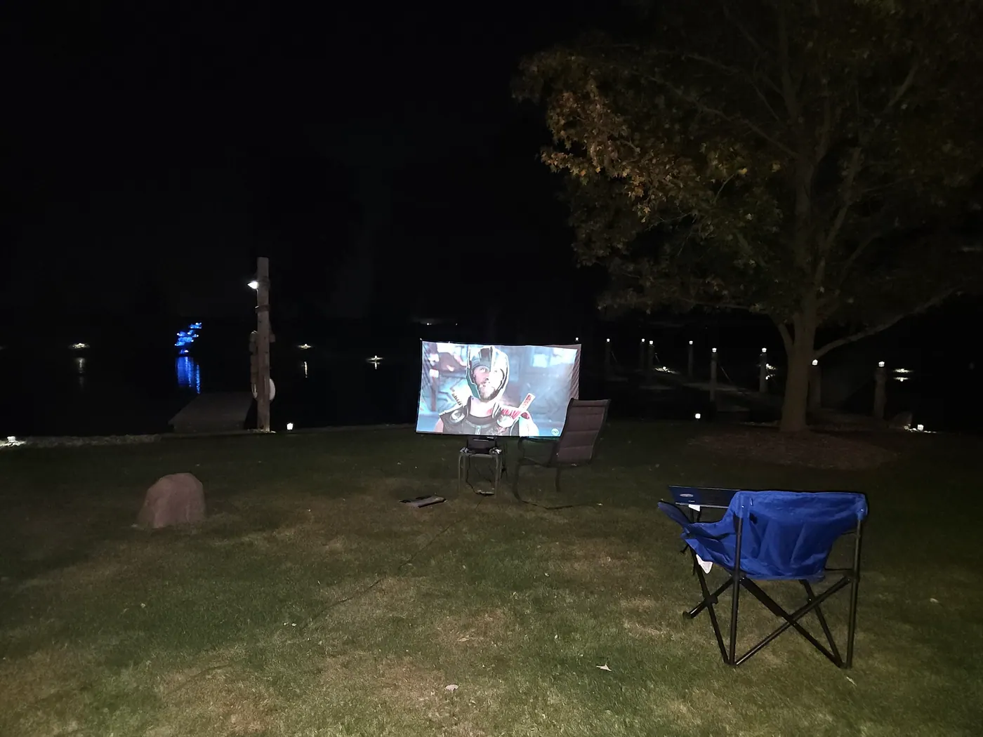 An outdoor movie projected on a screen set up on the lawn beside the pond after dark.